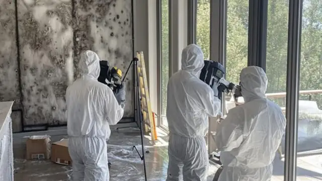 Three workers in protective suits setting up equipment in a large room with mold-stained walls