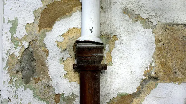Flaking plaster and discoloration around a rusted pipe on a damaged wall